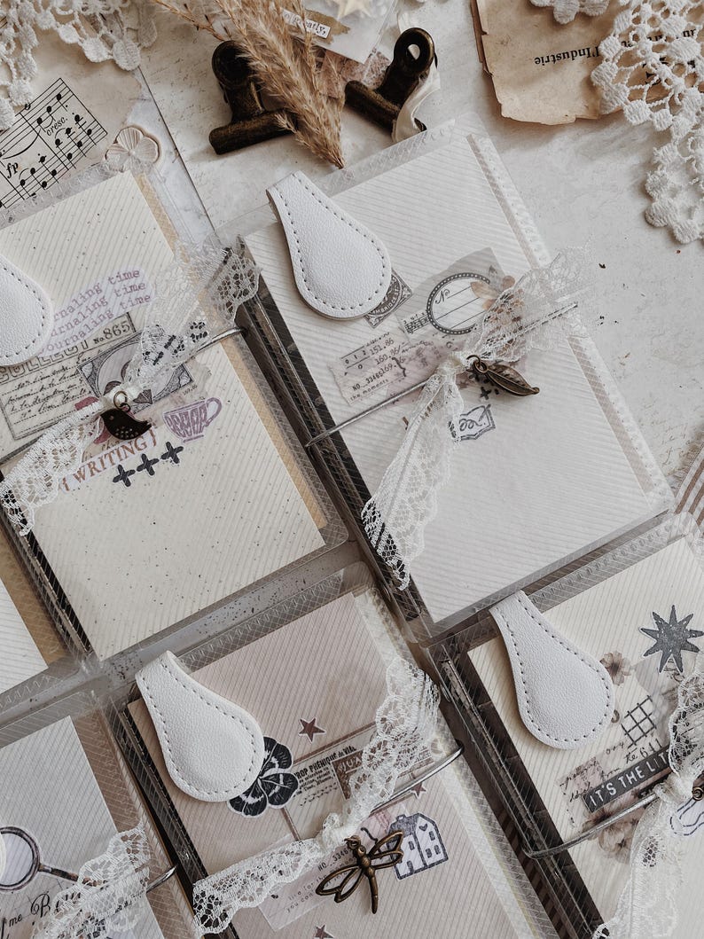 May include: A collection of rectangular journals featuring cream-colored paper with diagonal lines, secured by white teardrop-shaped magnetic clasps. Each journal is wrapped in white lace ribbon and adorned with metal charms and vintage-style stamps.