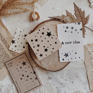 Pode incluir: Uma coleção de artigos de papelaria com tema celestial. Inclui blocos de madeira e cartões adornados com padrões de estrelas pretas. Um cartão diz "A new idea" e outro "Wish List". Os itens estão dispostos em uma superfície rústica e texturizada.