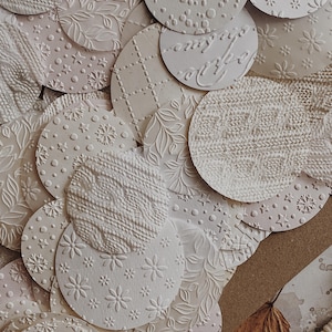 May include: A collection of off-white, textured paper circles with various embossed patterns. Designs include floral, knit, dot, and geometric motifs. The circles are layered, creating a tactile and visually appealing arrangement, suitable for crafts or scrapbooking.