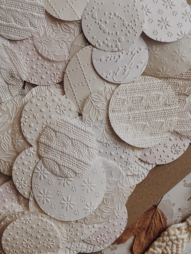 May include: A collection of off-white, textured paper circles with various embossed patterns. Designs include floral, knit, dot, and geometric motifs. The circles are layered, creating a tactile and visually appealing arrangement, suitable for crafts or scrapbooking.