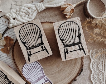 Pode incluir: Dois carimbos de madeira com um desenho de cadeira preta, juntamente com recortes de papel em forma de cadeira. Os carimbos estão em uma fatia de madeira, com fita decorativa, flores secas e um elemento de macramê. O fundo inclui papel vintage.