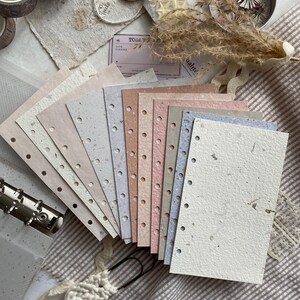 May include: A set of rectangular, multi-colored paper sheets with punched holes along one edge, featuring a textured, speckled finish in shades of cream, beige, pink, and blue, arranged in a fan shape next to a metal binder ring mechanism.