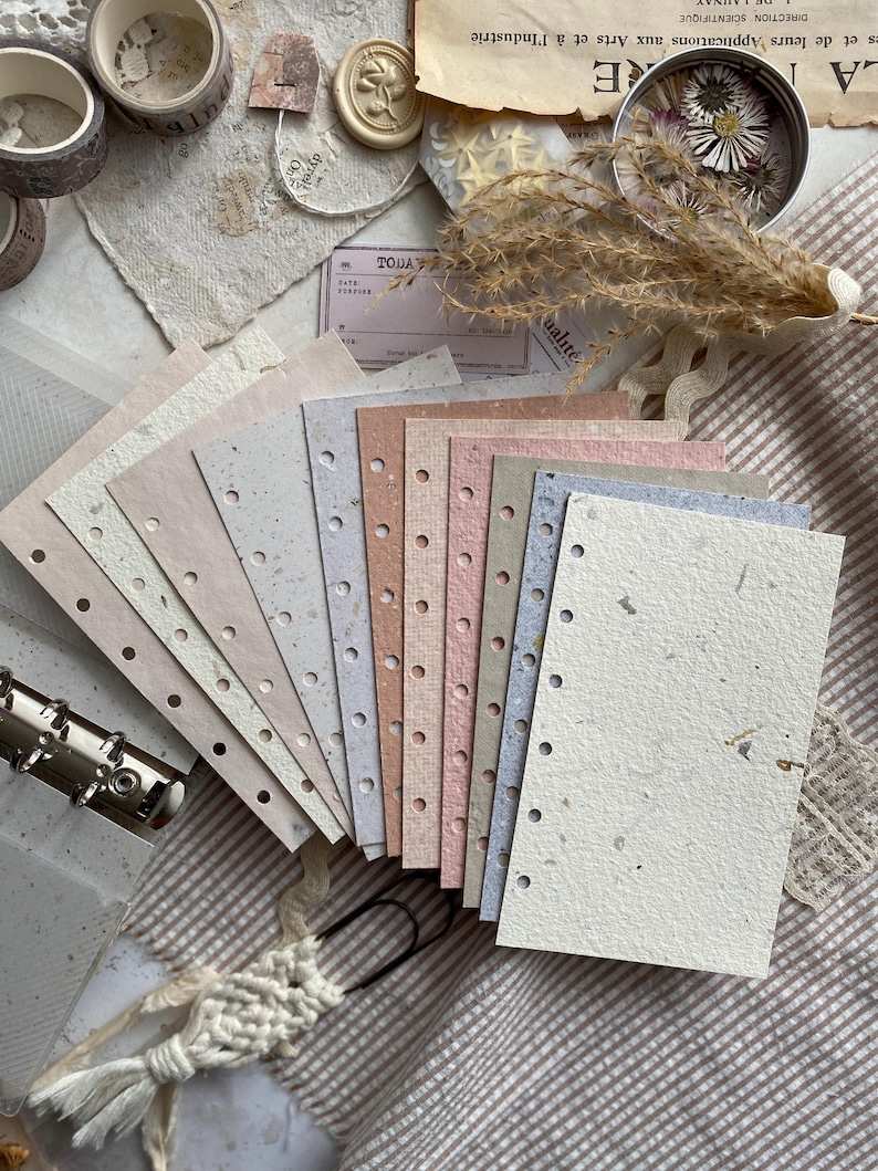 May include: A set of rectangular, multi-colored paper sheets with punched holes along one edge, featuring a textured, speckled finish in shades of cream, beige, pink, and blue, arranged in a fan shape next to a metal binder ring mechanism.
