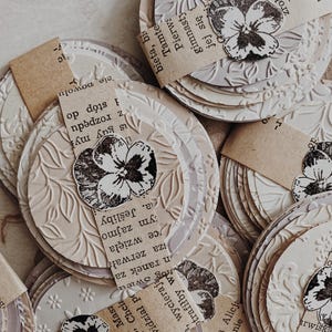 May include: A collection of layered, circular paper embellishments with embossed floral patterns. Each circle features a stamped black and white pansy and a small piece of paper with text. The phrase "HAVE A NICE DAY" is visible at the top.