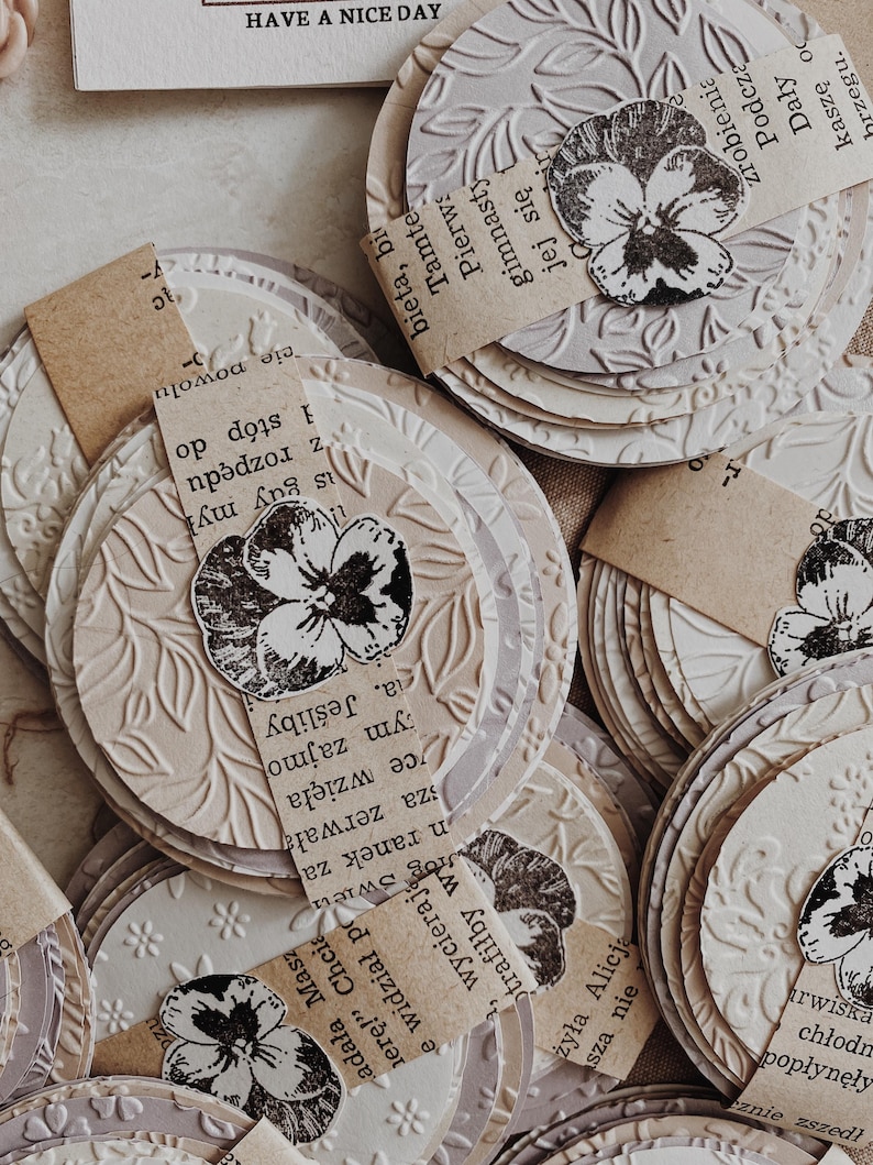 May include: A collection of layered, circular paper embellishments with embossed floral patterns. Each circle features a stamped black and white pansy and a small piece of paper with text. The phrase "HAVE A NICE DAY" is visible at the top.