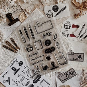 Pode incluir: Uma folha de carimbos de silicone transparente com vários ícones de papelaria, incluindo canetas-tinteiro, tinteiros, cadernos, clipes, óculos e balões de fala com texto como #penaddict, #stationerylover, #inkcollector e hello new color.