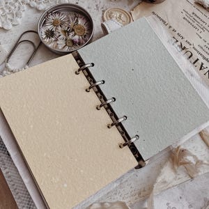 May include: A ring-bound planner featuring two distinct pages: one in a soft cream color and the other in a muted sage green. The planner has a metal six-ring mechanism holding the textured paper sheets in place, with visible flecks throughout.