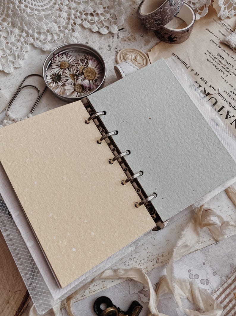 May include: A ring-bound planner featuring two distinct pages: one in a soft cream color and the other in a muted sage green. The planner has a metal six-ring mechanism holding the textured paper sheets in place, with visible flecks throughout.
