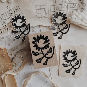 May include: A rectangular wooden rubber stamp featuring a black ink design of a bird centered within a flower blossom. The stamp is surrounded by three paper impressions of the same bird and flower motif, showcasing the detailed black ink illustration.