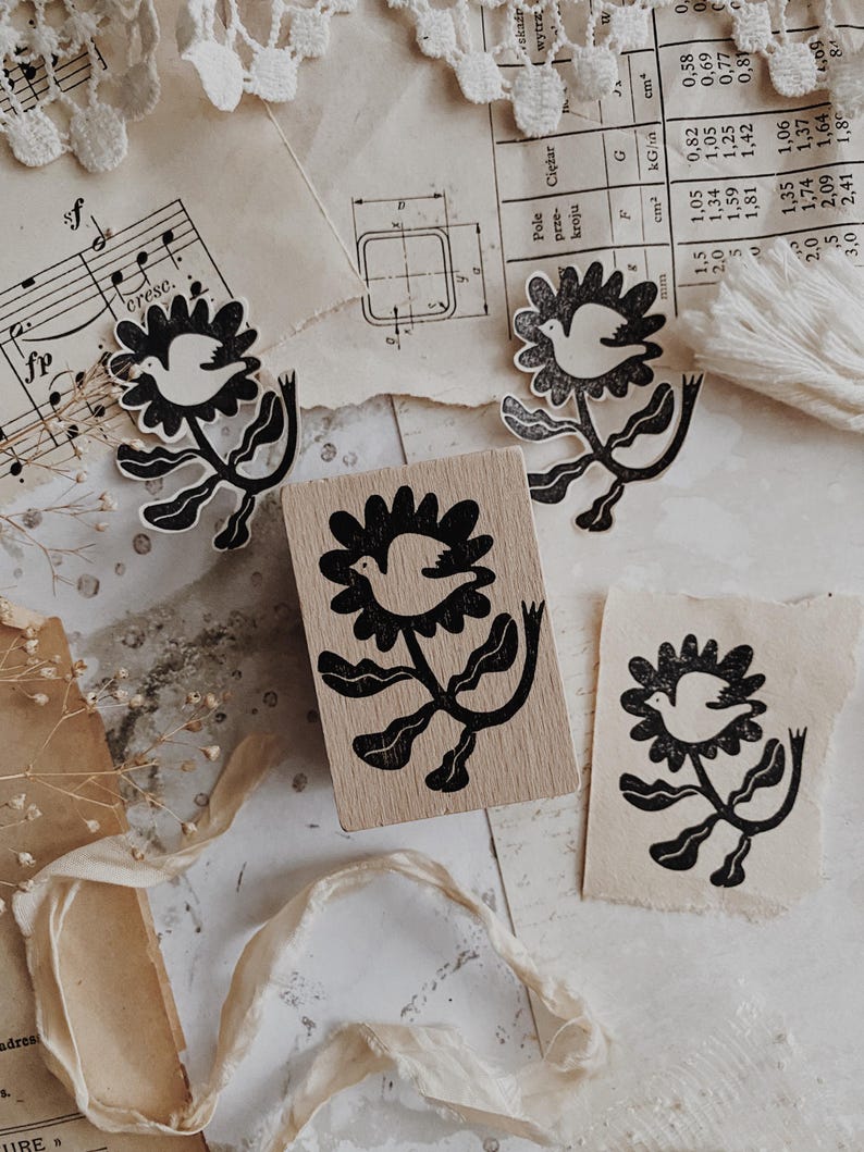 May include: A rectangular wooden rubber stamp featuring a black ink design of a bird centered within a flower blossom. The stamp is surrounded by three paper impressions of the same bird and flower motif, showcasing the detailed black ink illustration.