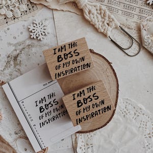 May include: Two rectangular wooden rubber stamps featuring the black printed text I AM THE BOSS OF MY OWN INSPIRATION. One stamp rests on a circular wood slice, while the other is placed above it, accompanied by a printed paper schedule.