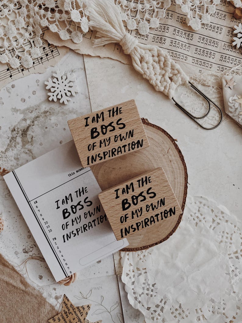 May include: Two rectangular wooden rubber stamps featuring the black printed text I AM THE BOSS OF MY OWN INSPIRATION. One stamp rests on a circular wood slice, while the other is placed above it, accompanied by a printed paper schedule.