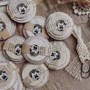 May include: A collection of handmade paper coasters with embossed floral designs and attached paper accents. Each coaster features a pressed flower image and is arranged on a neutral-toned surface, creating a vintage aesthetic.