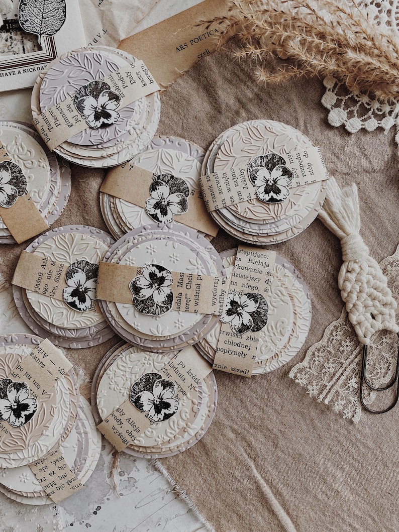 May include: A collection of handmade paper coasters with embossed floral designs and attached paper accents. Each coaster features a pressed flower image and is arranged on a neutral-toned surface, creating a vintage aesthetic.