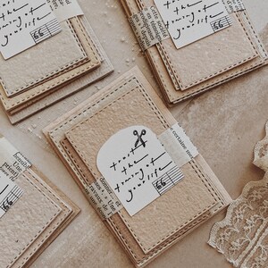May include: Several beige paper cards with stitched edges are arranged on a light surface. Each card has a small, arched tag with the text "trust the timing of your life" and a scissors graphic. Some cards have musical notes.