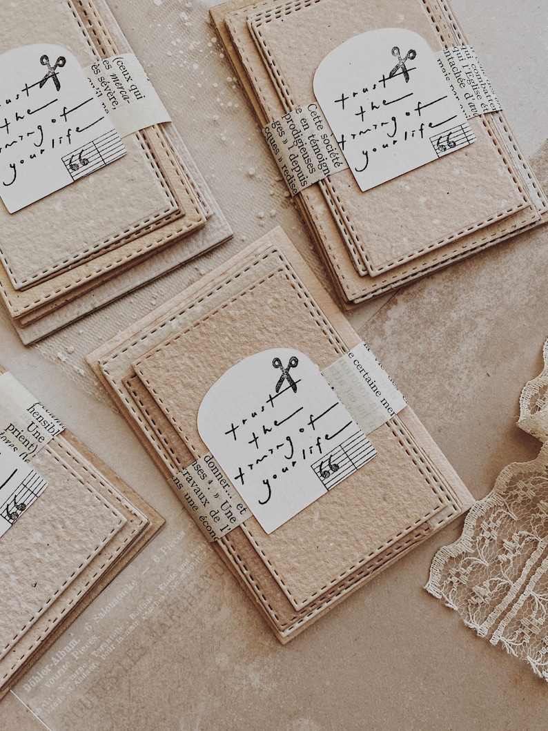 May include: Several beige paper cards with stitched edges are arranged on a light surface. Each card has a small, arched tag with the text "trust the timing of your life" and a scissors graphic. Some cards have musical notes.