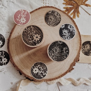 Peut inclure: Ensemble de tampons ronds en bois avec des motifs floraux. Les tampons, avec des motifs floraux blancs sur fond noir et rose, sont disposés sur une tranche de bois. Des feuilles séchées et du papier avec des croquis floraux complètent la composition.