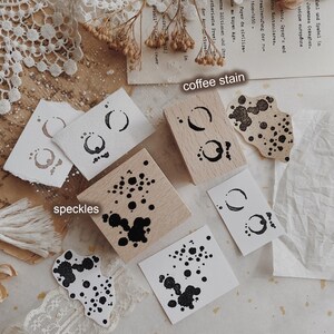 May include: Two square wooden stamps featuring black ink designs. One stamp displays a coffee stain pattern, and the other features a speckles design. The stamps are shown with corresponding black ink impressions on white paper, labeled coffee stain and speckles.