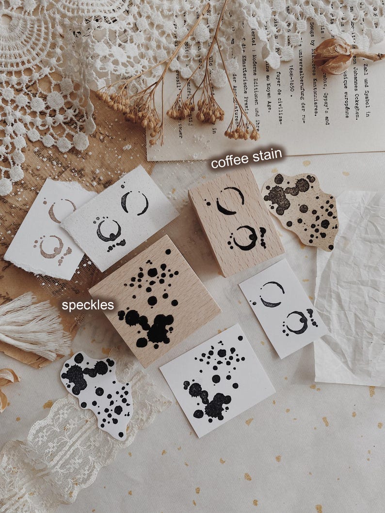 May include: Two square wooden stamps featuring black ink designs. One stamp displays a coffee stain pattern, and the other features a speckles design. The stamps are shown with corresponding black ink impressions on white paper, labeled coffee stain and speckles.