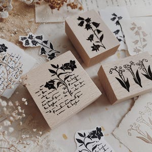 May include: Three rectangular wooden stamps featuring black ink designs. One stamp displays a bellflower with cursive script, another shows a single bellflower stem, and the third features a cluster of lily of the valley flowers with leaves.