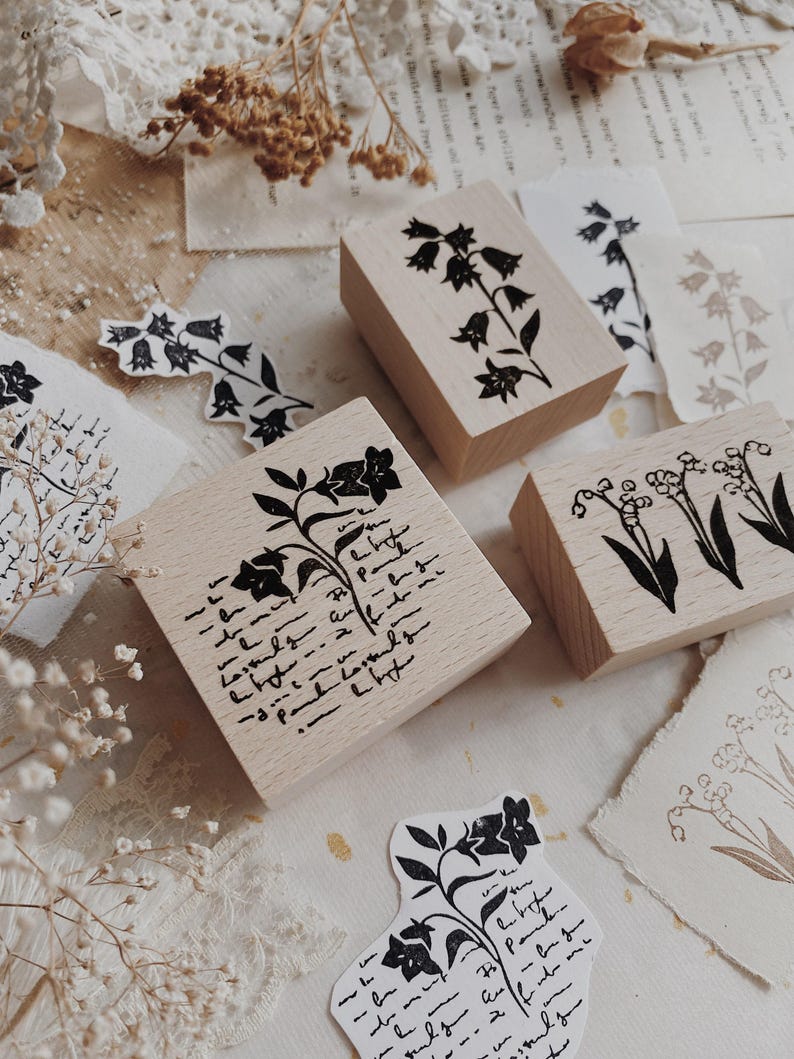 May include: Three rectangular wooden stamps featuring black ink designs. One stamp displays a bellflower with cursive script, another shows a single bellflower stem, and the third features a cluster of lily of the valley flowers with leaves.