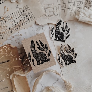 May include: A rectangular wooden rubber stamp and two die-cut paper stickers, each featuring a black ink illustration of a rabbit surrounded by leaves. The stamp has a light wood grain finish, while the stickers are off-white with black ink.