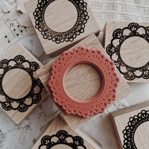 May include: Five square wooden stamps with light brown grain. Four stamps feature black ink impressions of circular, scalloped lace-like frames. One central stamp displays a raised, terracotta-colored rubber die with a matching circular lace pattern.