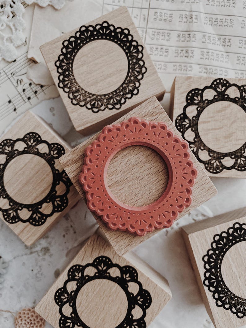 May include: Five square wooden stamps with light brown grain. Four stamps feature black ink impressions of circular, scalloped lace-like frames. One central stamp displays a raised, terracotta-colored rubber die with a matching circular lace pattern.