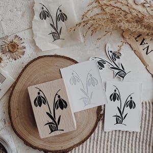 May include: A rectangular wooden rubber stamp featuring a black ink illustration of two snowdrop flowers with leaves. The stamp is accompanied by several white paper prints displaying the same floral design in black ink.