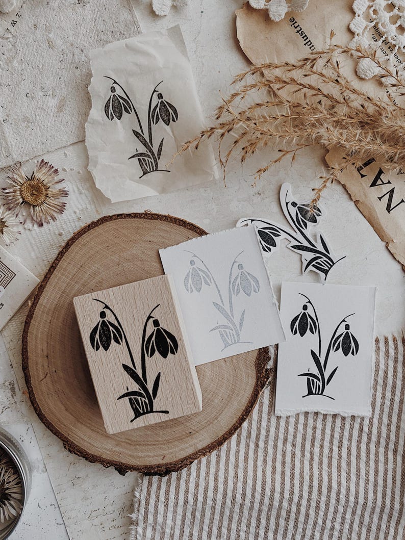 May include: A rectangular wooden rubber stamp featuring a black ink illustration of two snowdrop flowers with leaves. The stamp is accompanied by several white paper prints displaying the same floral design in black ink.