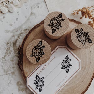 Puede incluir: Sellos de madera con un diseño de rosa y un boleto con el texto "TICKET SWEET TIME" y "CHERISH THE TIME". Los sellos están dispuestos sobre una rodaja de madera, con papel y encaje vintage en el fondo.