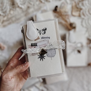 Può includere: Un piccolo taccuino con copertina in plastica trasparente e chiusura magnetica in similpelle bianca. Il fronte presenta un cartoncino color crema con grafiche nere stampate di una farfalla, una foglia, una stella e il testo planning e 1054.