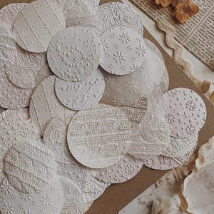 May include: A collection of white, textured paper circles with various embossed patterns, including floral, knit, and geometric designs. The circles are arranged on a brown surface with vintage paper and dried leaves.