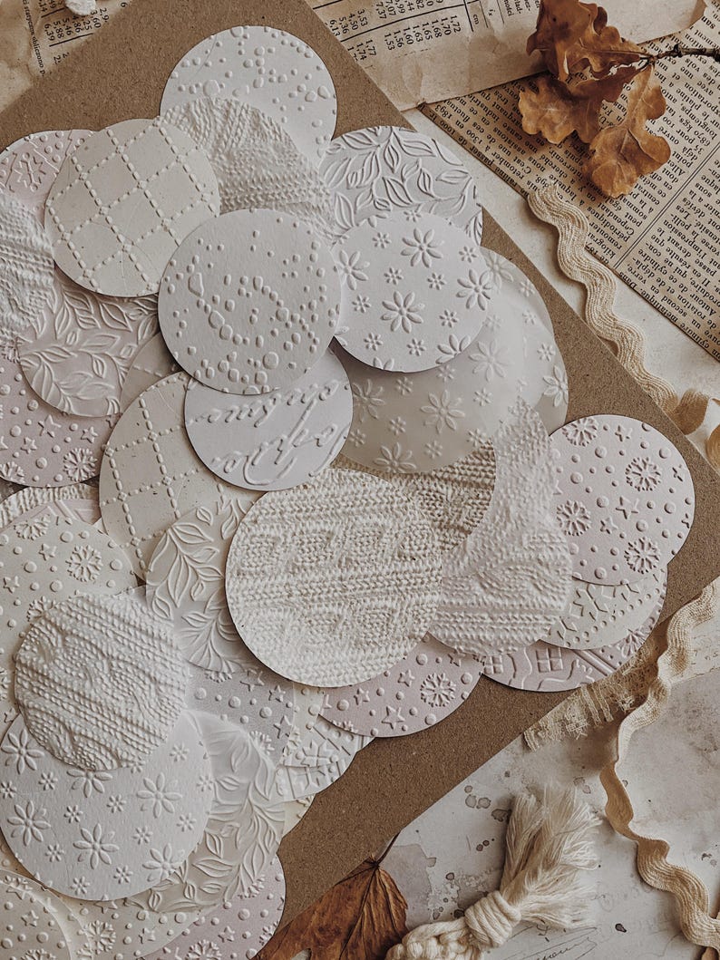 May include: A collection of white, textured paper circles with various embossed patterns, including floral, knit, and geometric designs. The circles are arranged on a brown surface with vintage paper and dried leaves.
