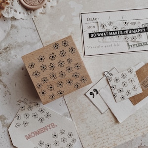May include: A wooden stamp featuring a pattern of small daisy motifs. The image includes stationery elements such as a wax seal, paper ephemera, and a card with the text "DO WHAT MAKES YOU HAPPY."