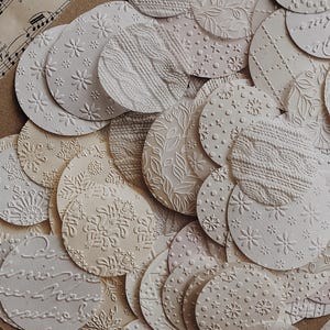 May include: A collection of off-white, embossed paper circles with various patterns, including floral, snowflake, and cable knit designs. The circles are arranged on a neutral-toned background, creating a textured and layered effect. Some circles have script text.