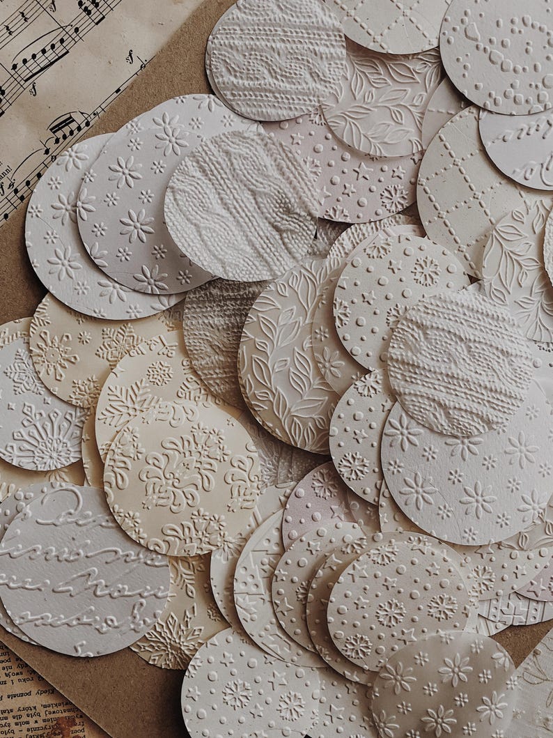 May include: A collection of off-white, embossed paper circles with various patterns, including floral, snowflake, and cable knit designs. The circles are arranged on a neutral-toned background, creating a textured and layered effect. Some circles have script text.