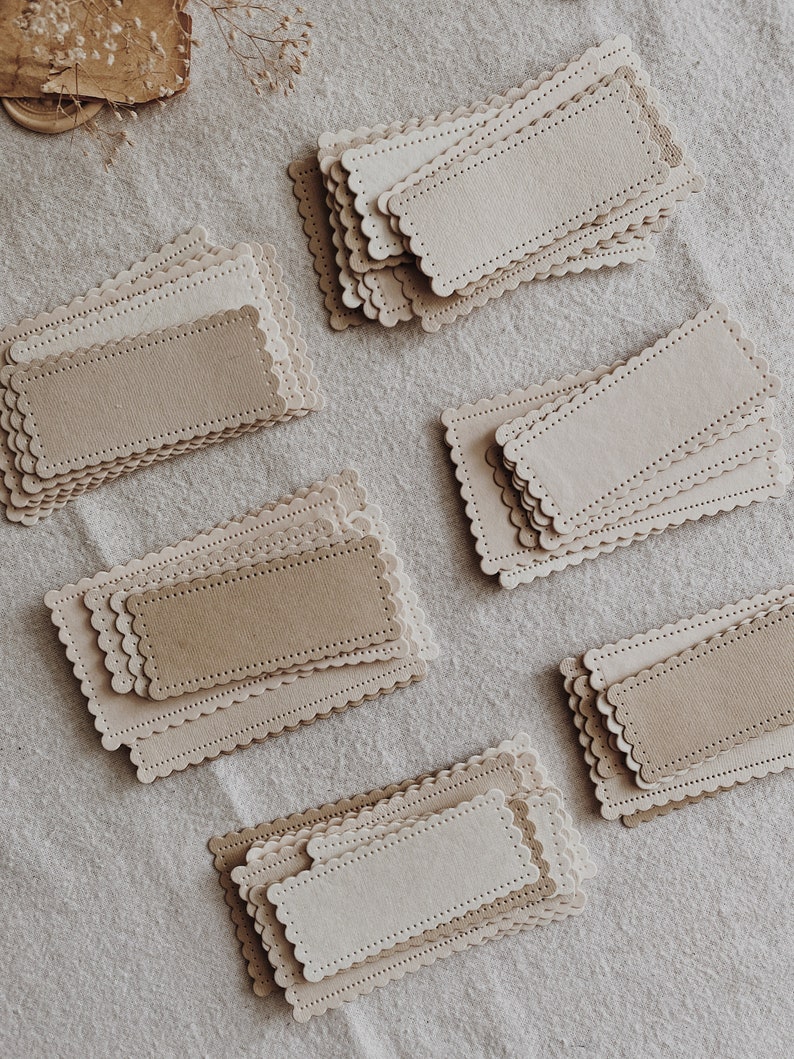 Puede incluir: Una colecci&oacute;n de etiquetas rectangulares de papel con bordes festoneados. Las etiquetas son de varios tonos de beige y marr&oacute;n, con una superficie texturizada. Cada etiqueta tiene un rect&aacute;ngulo m&aacute;s peque&ntilde;o y oscuro en el centro y una serie de peque&ntilde;os agujeros.