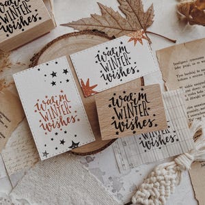 May include: A rectangular wooden rubber stamp featuring the text Warm Winter Wishes surrounded by small stars. The stamp is shown alongside two white paper cards stamped with the same design in black and copper ink, displaying the phrase clearly.