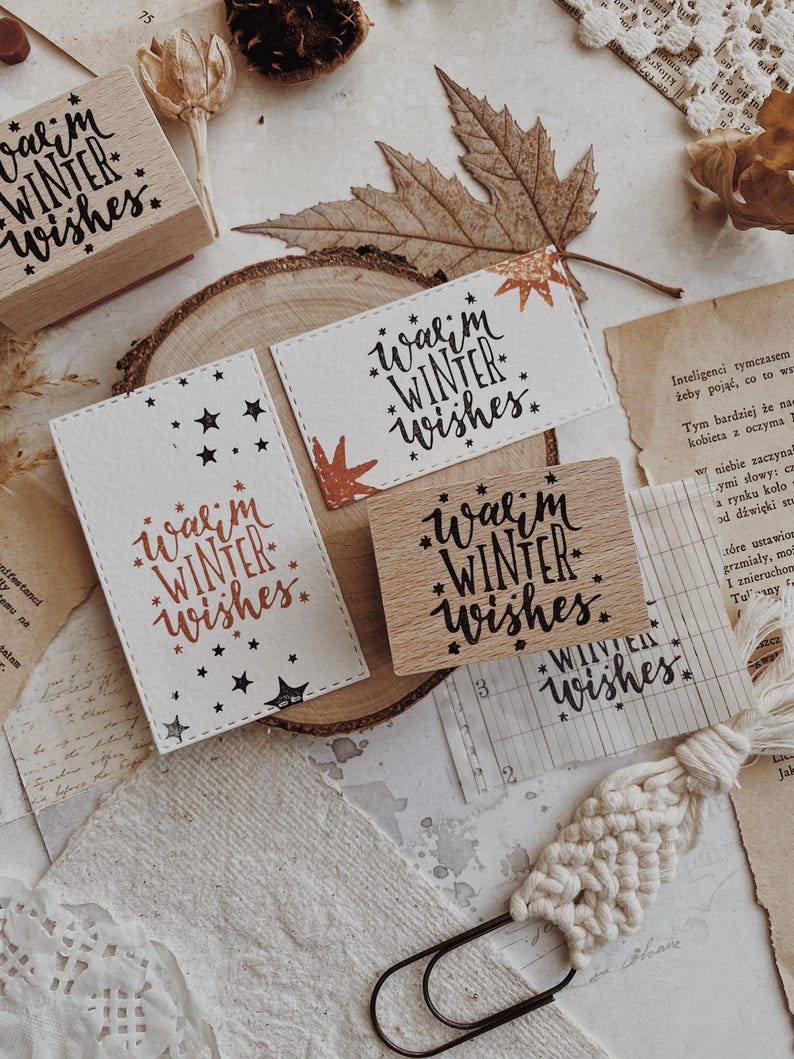 May include: A rectangular wooden rubber stamp featuring the text Warm Winter Wishes surrounded by small stars. The stamp is shown alongside two white paper cards stamped with the same design in black and copper ink, displaying the phrase clearly.