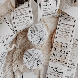 Puede incluir: Dos rollos de cinta washi etiquetados como albumlovehandmade 40mm/5m ALW2613, rodeados de tiras de papel de estilo vintage con texto impreso, notación musical y gráficos, junto a un marcapáginas de ganchillo color crema con un clip metálico.