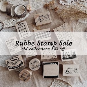 May include: A collection of wooden rubber stamps featuring various designs including a swan, a window, a forest with a deer, and text such as I like big CUPS and Enjoy the AUTUMN. The stamps are arranged alongside rolls of patterned tape.