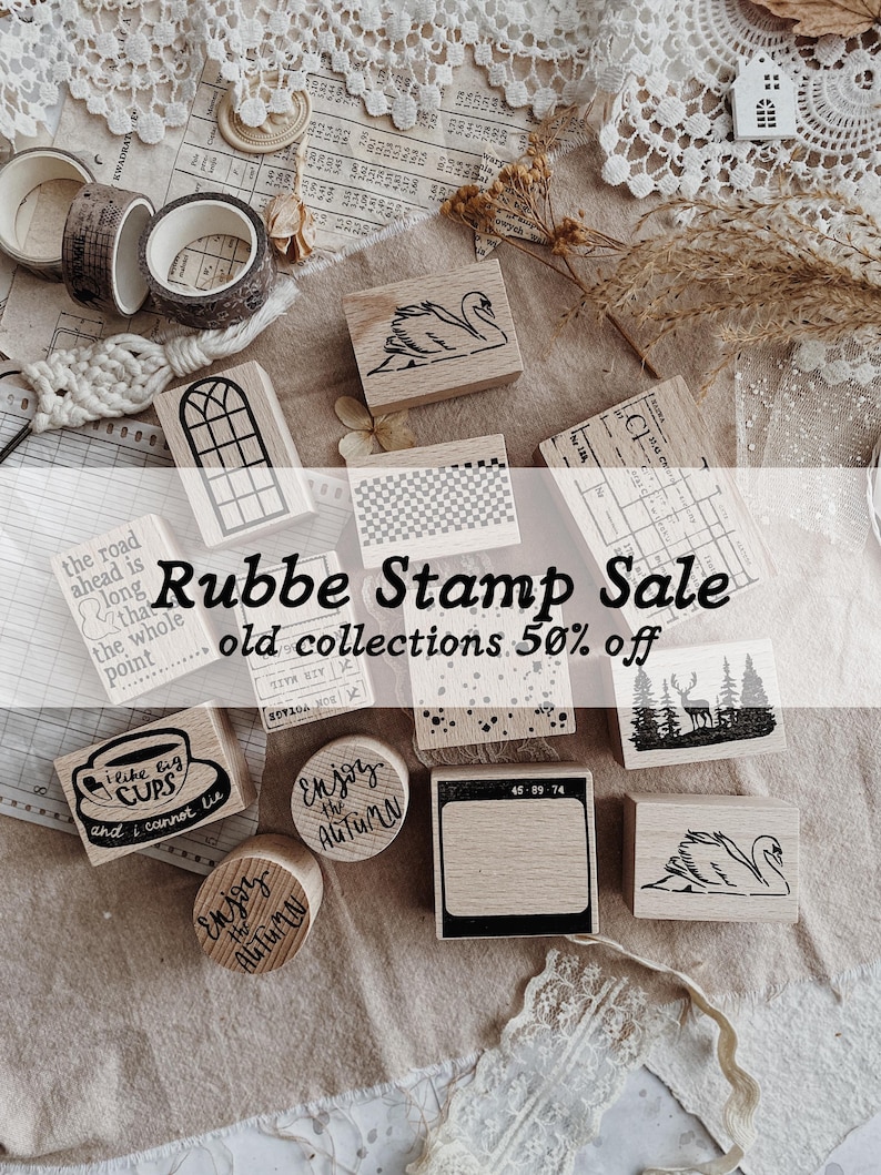 May include: A collection of wooden rubber stamps featuring various designs including a swan, a window, a forest with a deer, and text such as I like big CUPS and Enjoy the AUTUMN. The stamps are arranged alongside rolls of patterned tape.