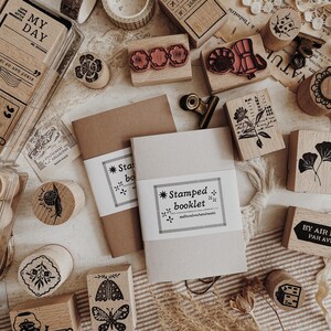 May include: A collection of wooden stamps with various designs, including flowers, butterflies, and text, alongside notebooks and decorative tape. The stamps feature ink impressions in red and black. The text on the notebooks reads "Stamped booklet".