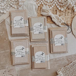 May include: A collection of handmade paper cards with stitched edges and a decorative paper band. Each card features the text "toast the turning of your life" and a musical note graphic. The cards are beige and cream colored.