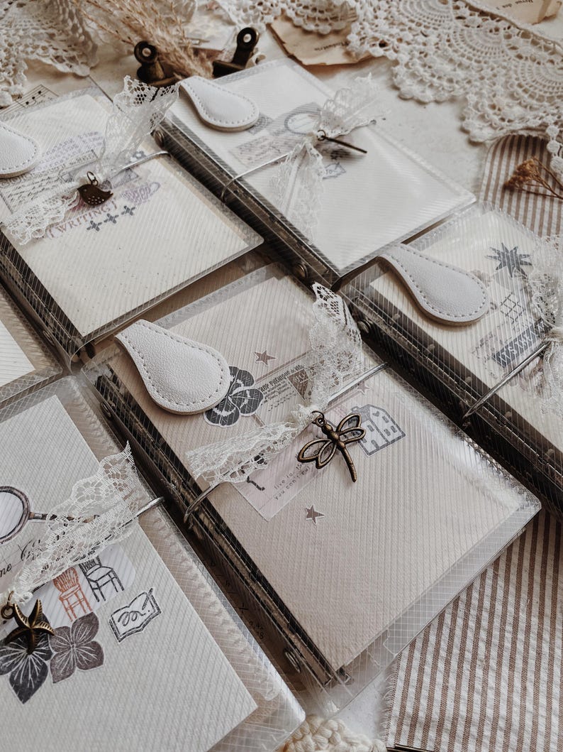 May include: A collection of rectangular notebooks encased in clear plastic covers, each featuring a white faux leather tab closure, white lace ribbon, and a small metal charm such as a dragonfly or bird, with printed illustrations on the paper.