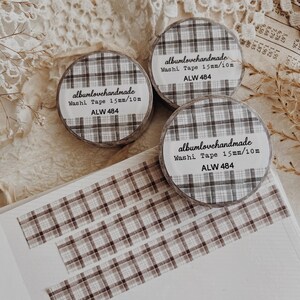 Può includere: Tre rotoli di washi tape con motivo a quadri grigio e marrone. Ogni rotolo ha un'etichetta con la scritta "albumlovehandmade Washi Tape 15mm/10m ALW 484". Strisce dello stesso nastro sono su una superficie bianca.