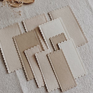 Puede incluir: Una colecci&oacute;n de etiquetas rectangulares de papel en varios tonos neutros, incluyendo beige, crema y topo. Cada etiqueta tiene un borde festoneado y una l&iacute;nea de peque&ntilde;as perforaciones. Las etiquetas est&aacute;n dispuestas sobre una superficie texturizada de color claro.