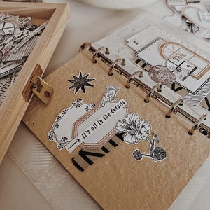 May include: A ring-bound journal with tan paper pages decorated with black and white stickers, including a star, a flower, and a label with the text "it's all in the details" featuring a pointing hand icon and a small window illustration.