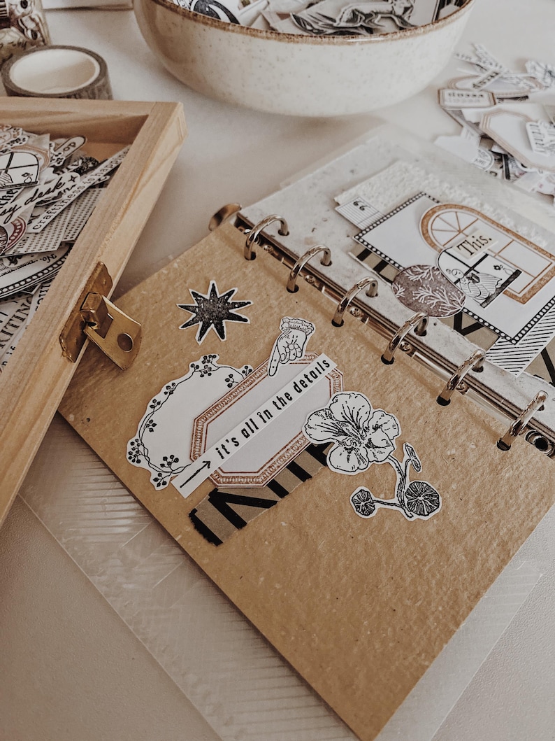May include: A ring-bound journal with tan paper pages decorated with black and white stickers, including a star, a flower, and a label with the text "it's all in the details" featuring a pointing hand icon and a small window illustration.